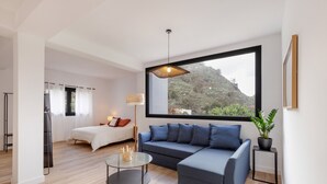 Een moderne woonkamer met een bank, een glazen salontafel, een bed en een groot raam met uitzicht op groen.