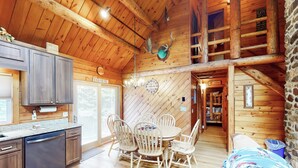 Interior rústico de una cabaña con mesa y sillas de madera, cocina con lavavajillas y pared de piedra.