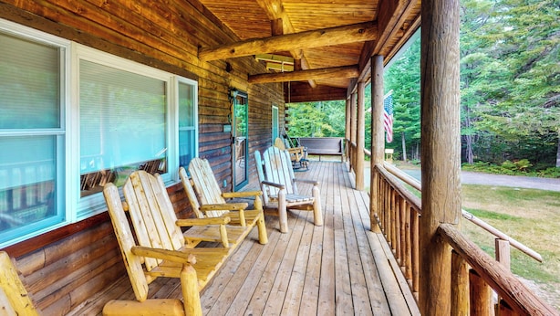 Un porche de madera con sillas Adirondack, una mesa y vistas a un bosque.
