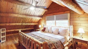 Un dormitorio de cabaña de madera con una cama, dos mesitas de noche con lámparas, una ventana con persianas y un ventilador de techo.