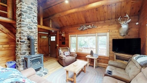 Interior de una cabaña de troncos con chimenea de piedra, mobiliario de madera, cabeza de venado montada y un televisor.