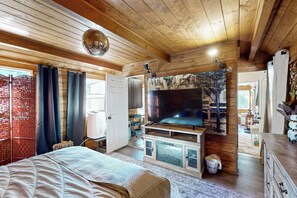 Une chambre confortable, aménagée dans le style d’un chalet, avec un grand lit, un téléviseur fixé au mur, des murs en bois et une fenêtre avec des rideaux.