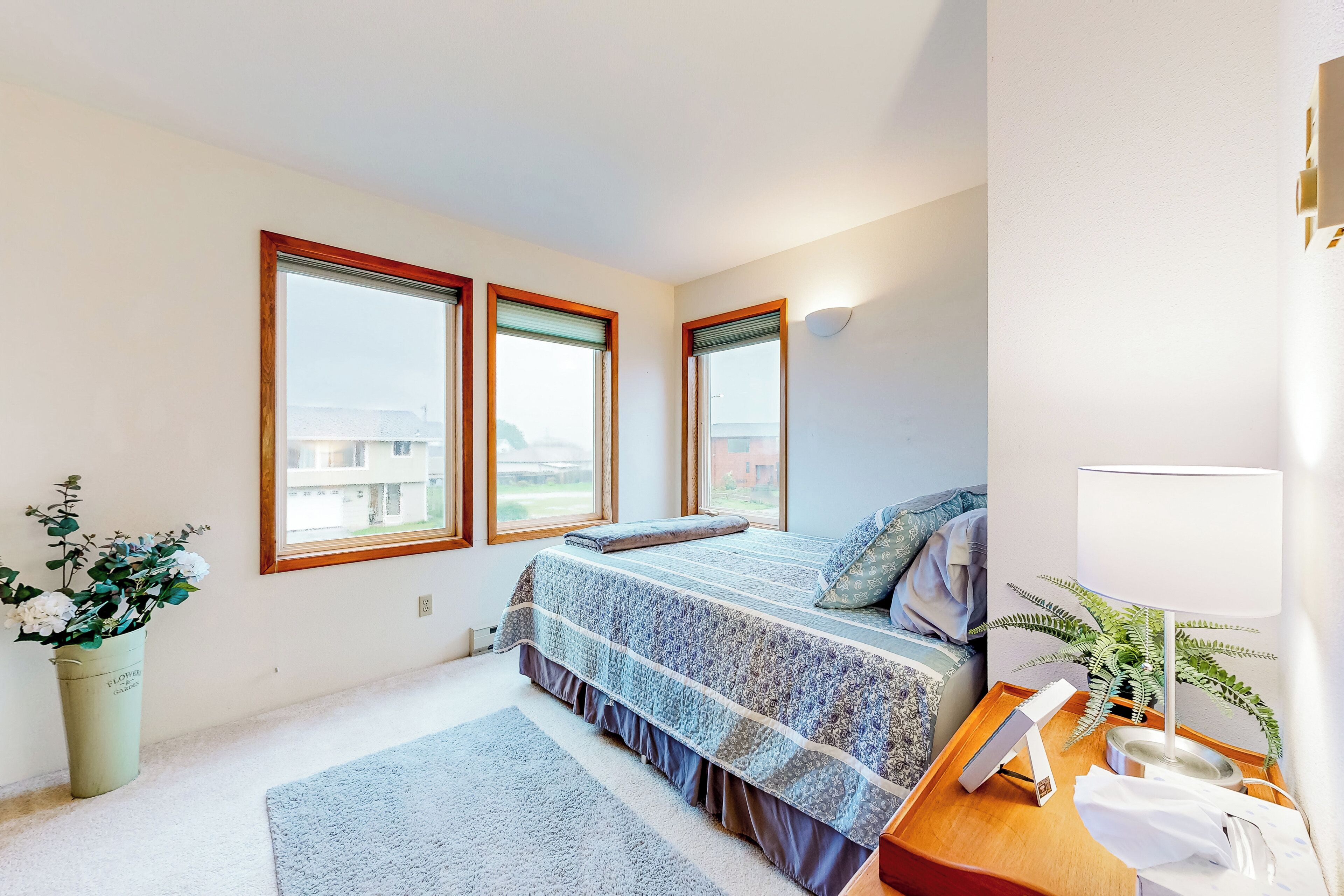 A bedroom with a large bed, a bedside table with a lamp, a potted plant, and a view of the outside through the windows.