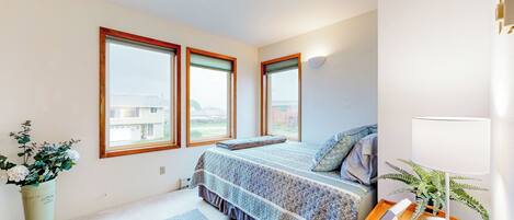 A bedroom with a large bed, a bedside table with a lamp, a potted plant, and a view of the outside through the windows.