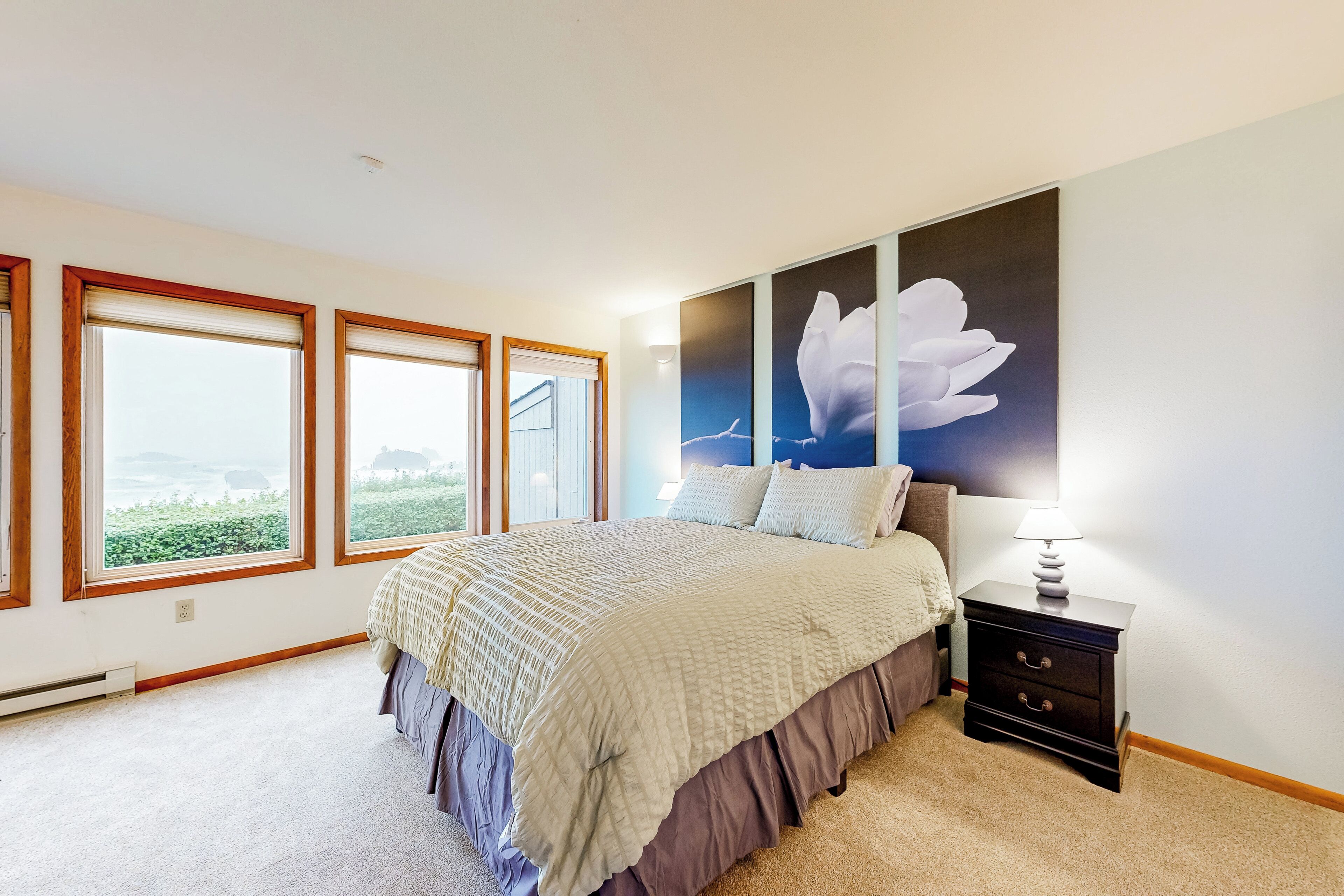 Un dormitorio con una cama grande, una mesita de noche y una lámpara fijada a la pared. Hay tres ventanas con marcos de madera y vistas al exterior con vegetación.