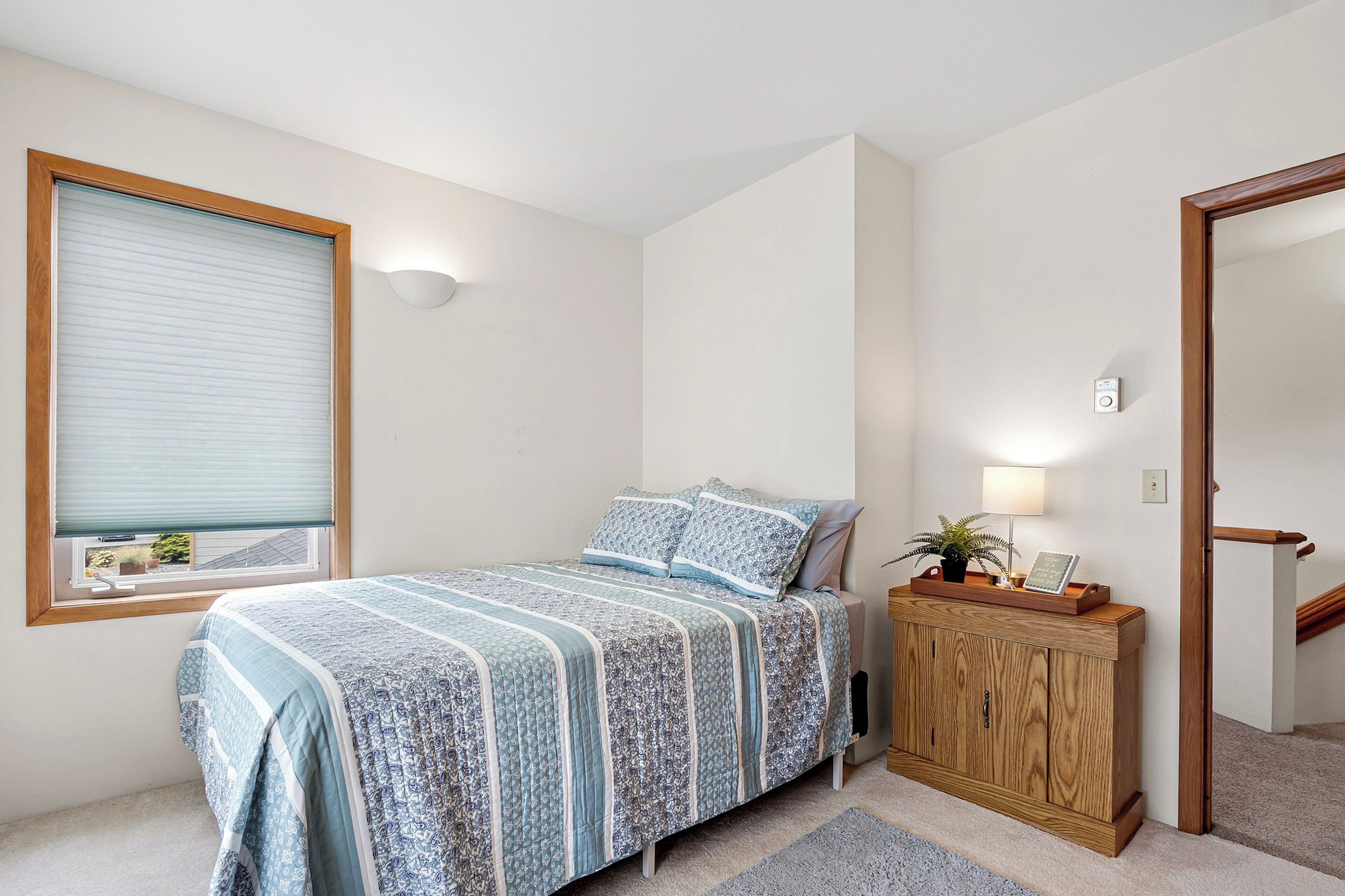 A bedroom with a bed, a nightstand, a lamp, a window with blinds, and a door to another room.