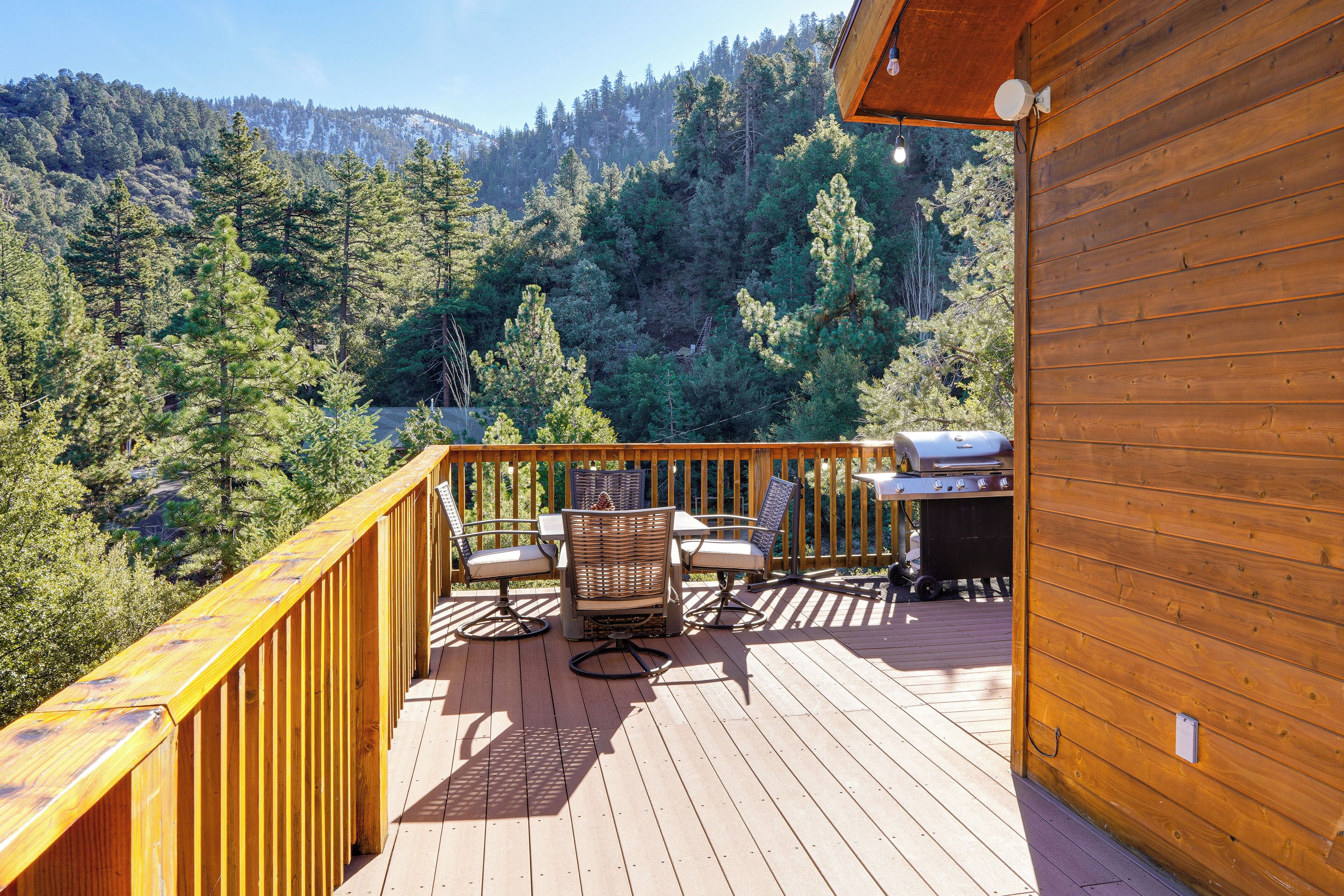 Una terraza de madera con mobiliario exterior, una parrilla y vistas a una zona montañosa boscosa.