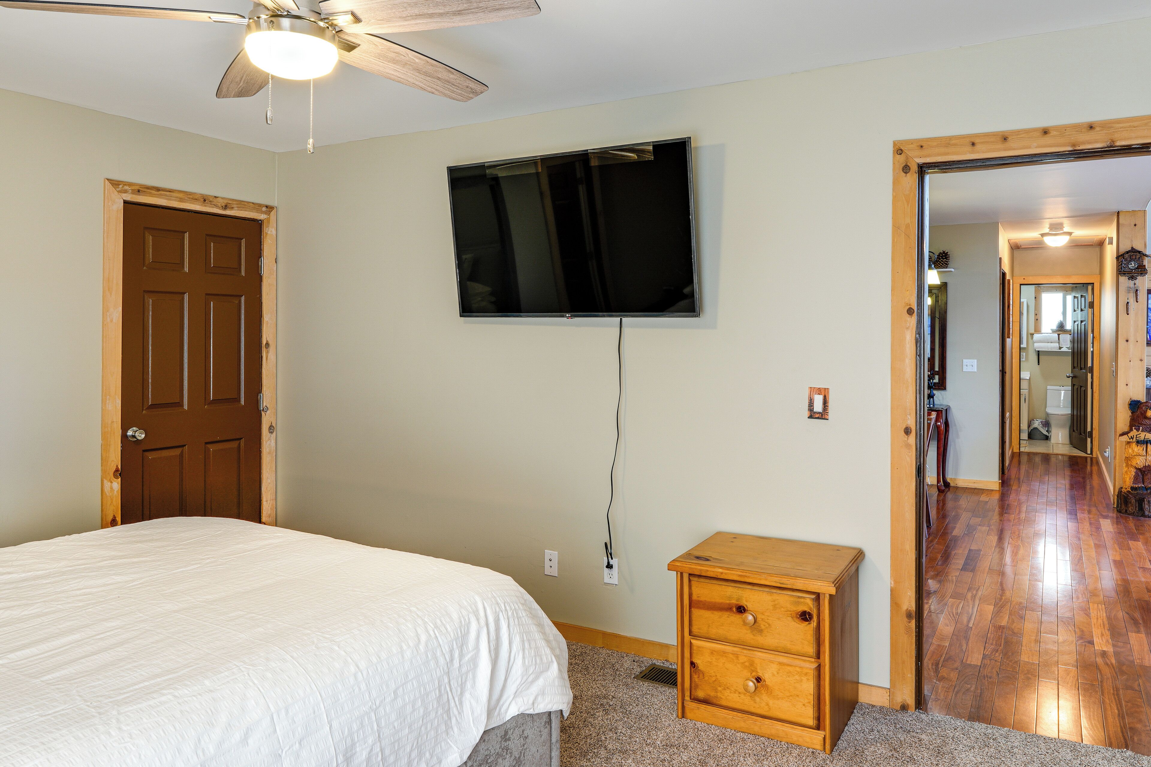 A bedroom with a bed, a TV mounted on the wall, a wooden nightstand, and a door leading to another room.