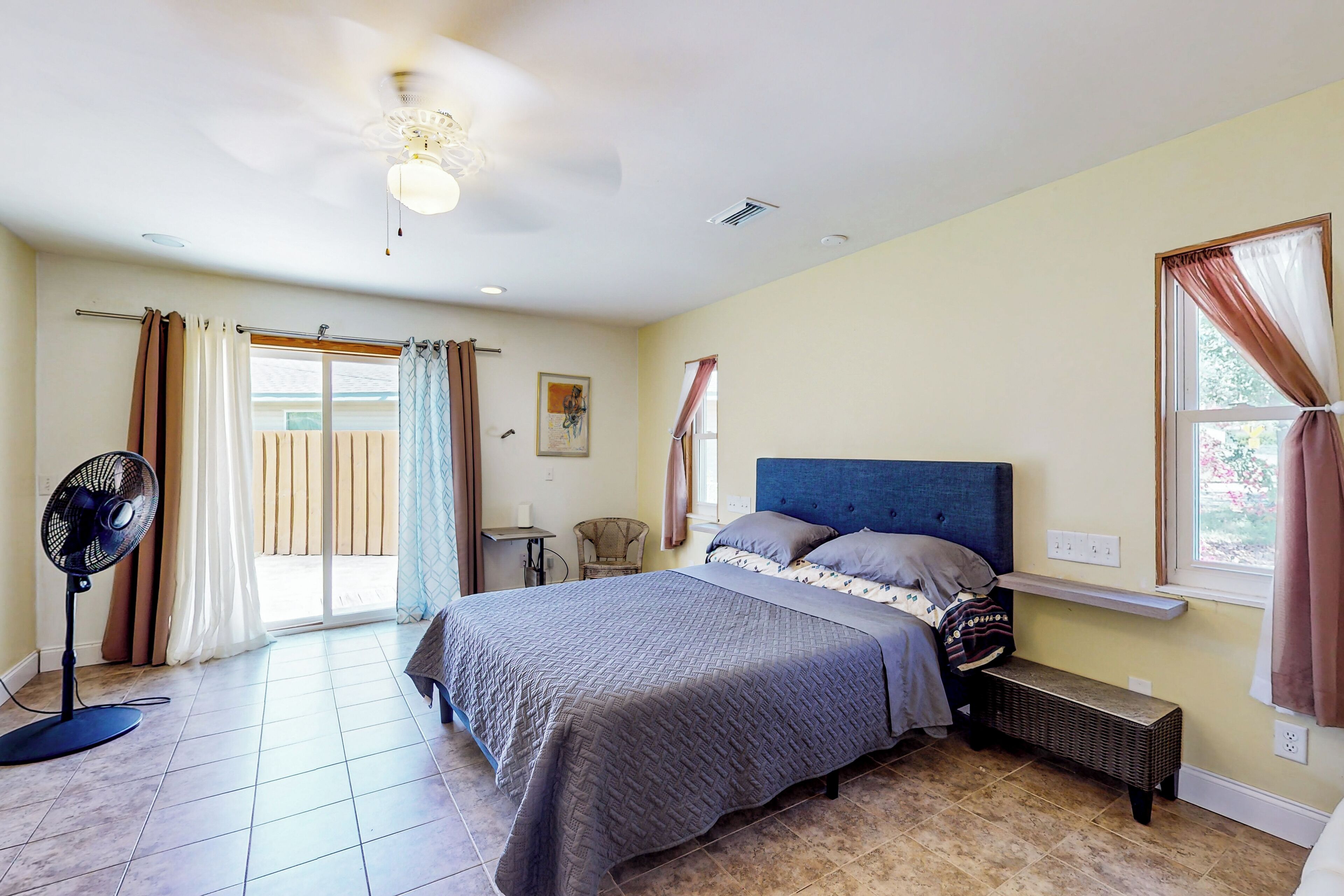 Quarto com cama, ventilador de teto, janela com cortinas e uma pequena mesa.