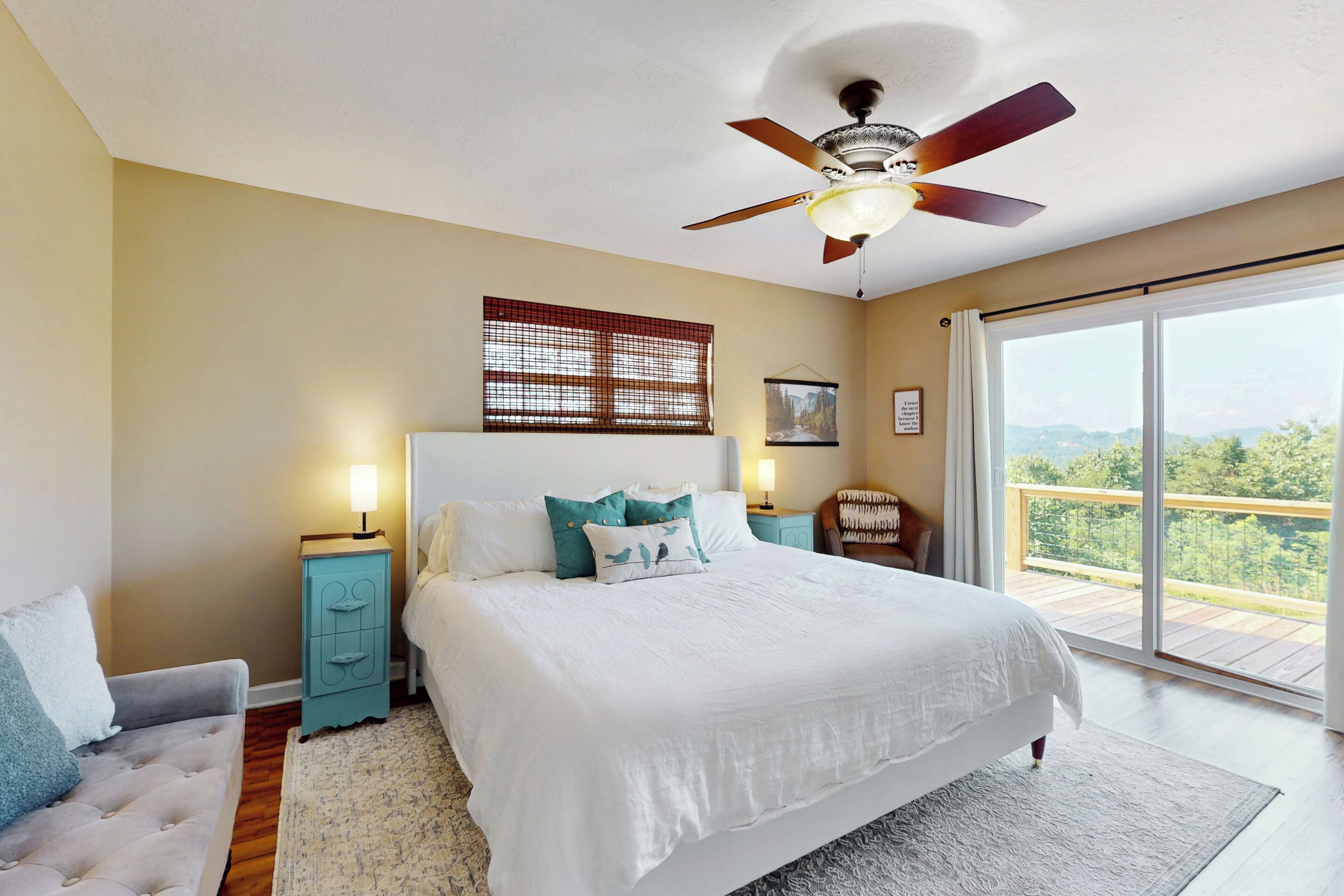A bedroom with a large bed, a ceiling fan, a window with blinds, a nightstand with a lamp, and a view of the outdoors.