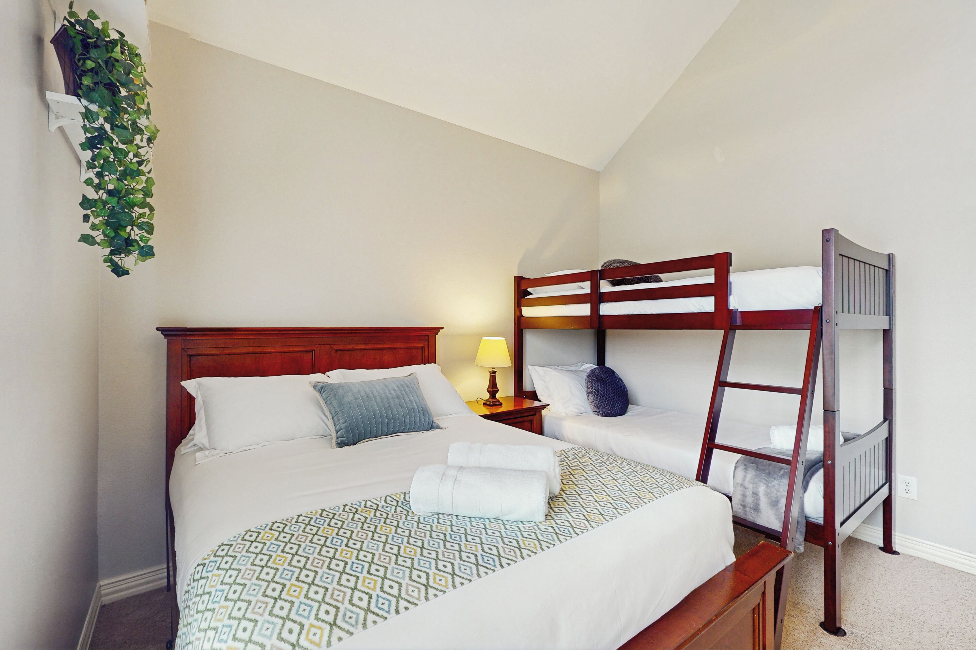 A bedroom with a large bed, a smaller bed, a wooden ladder, a nightstand with a lamp, and a hanging plant.