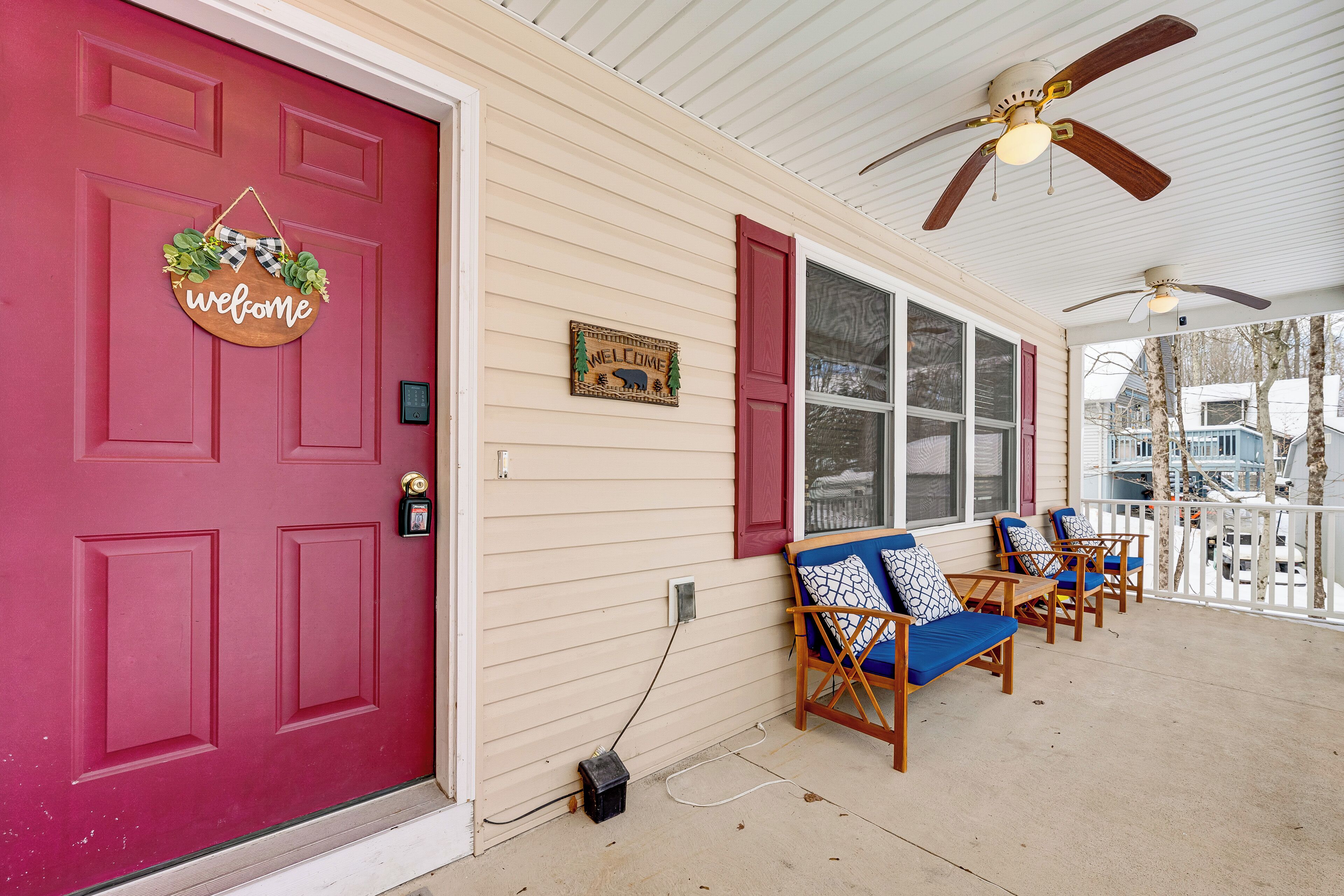 A porch with a red door, a welcome sign, and a 'Welcome' sign with a bear. There are blue chairs and a ceiling fan.