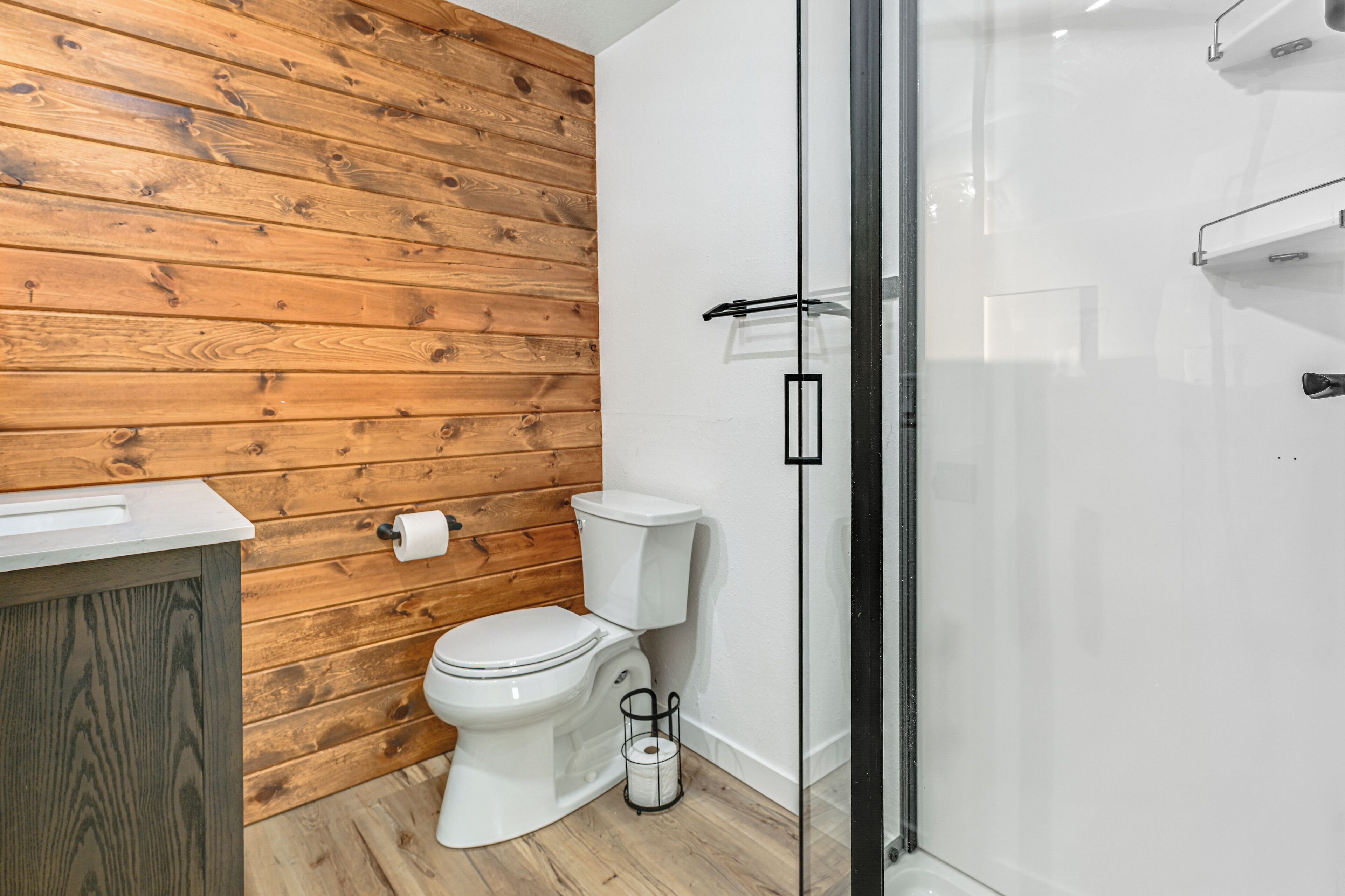A bathroom with a wooden wall, a white toilet, a glass shower enclosure, and a white sink.