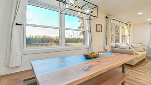 A modern living room with a wooden dining table, a sofa, and large windows offering a view of trees. - Riverfront Reset! Dells Cabin w/ Kayaks & Deck (Wisconsin Dells)