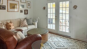 Een woonkamer met een bruine leren bank, een witte bank met gekleurde kussens, een rieten salontafel en een vloerkleed met een patroon. Er hangen afbeeldingen in lijsten aan de muur en een grote glazen deur met uitzicht.