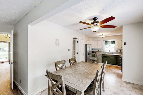 A dining room with a wooden table and chairs, a ceiling fan, and a kitchen in the background.