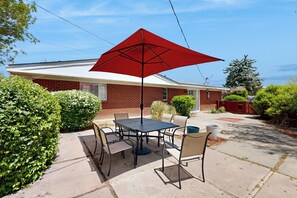 Una terrazza esterna con tavolo e sedie, un ombrello rosso e una casa in mattoni sullo sfondo.