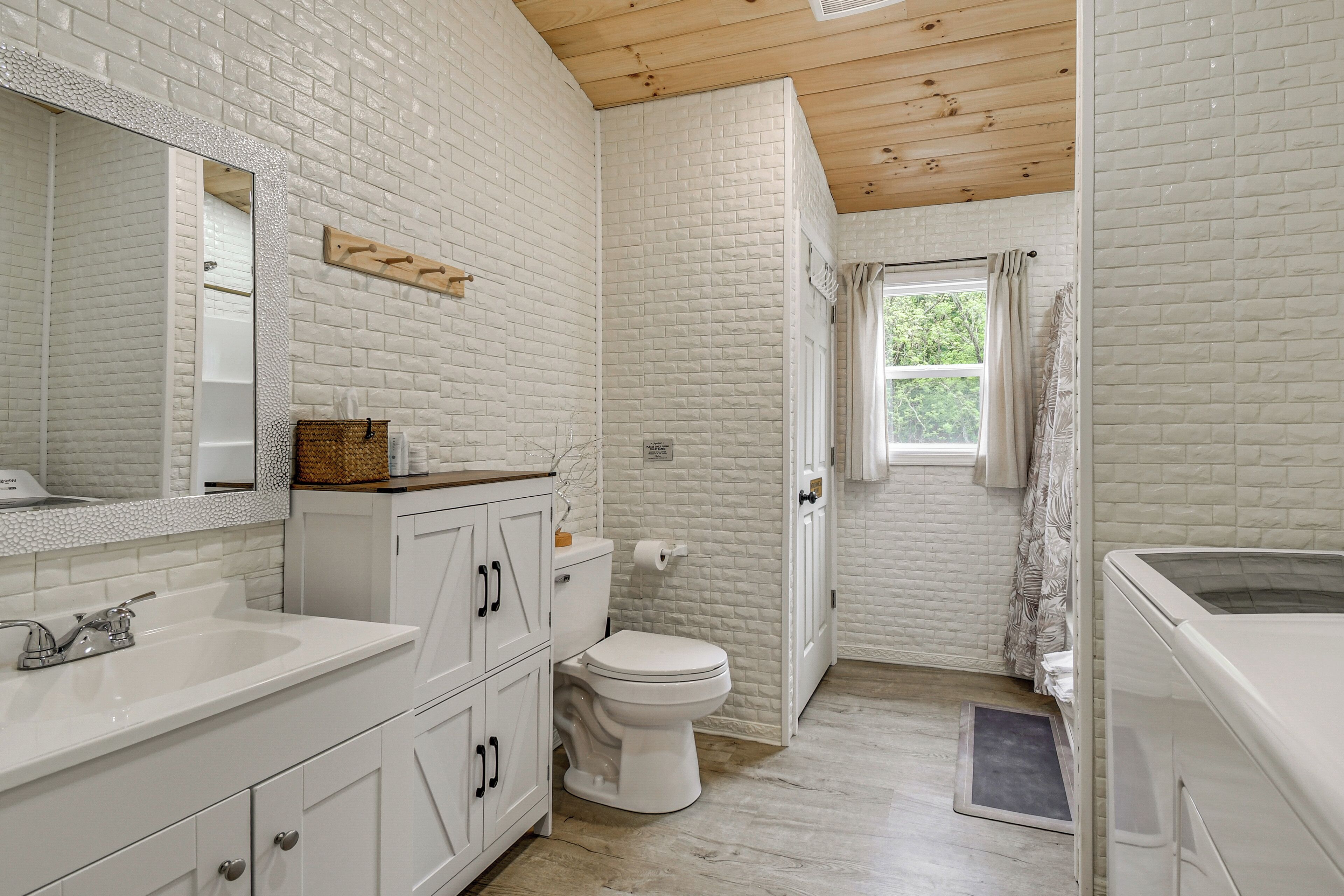 A bathroom with a white brick wall, wooden ceiling, a white sink, a toilet, and a window with curtains.