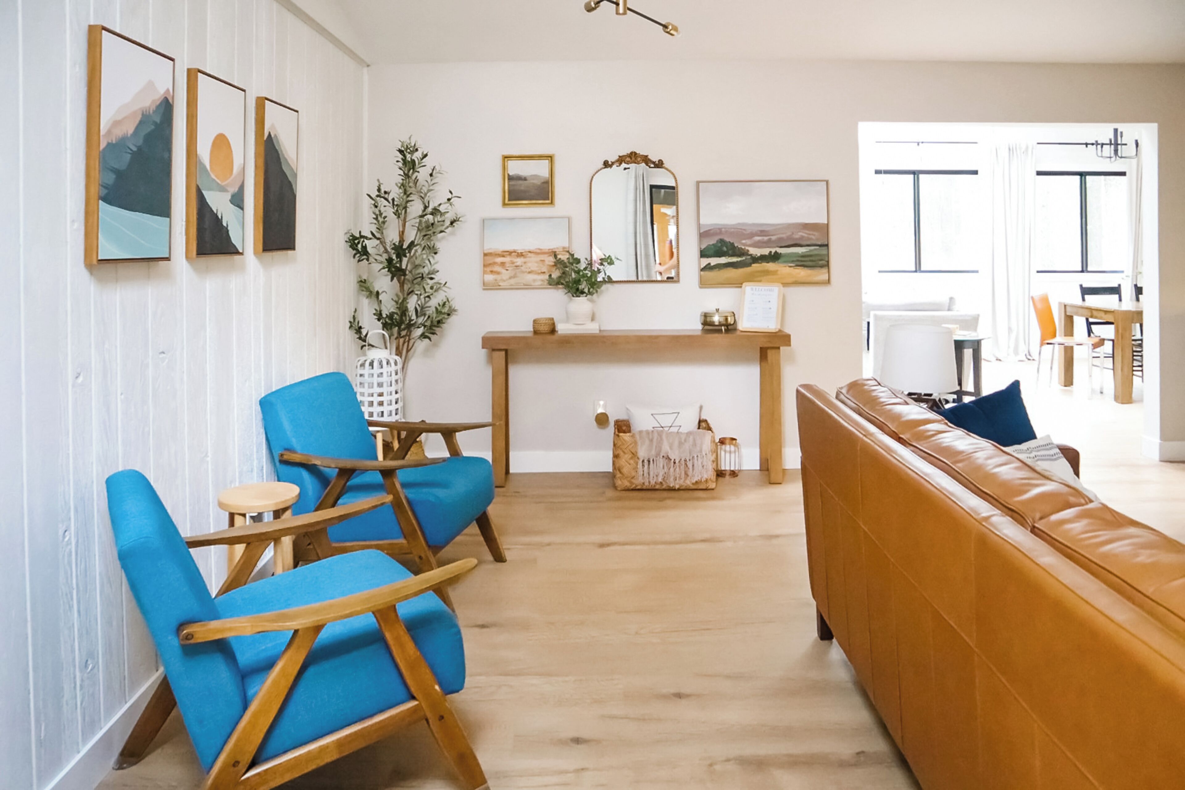 A living room with a wooden floor, a blue sofa, and a brown leather couch. There are two blue chairs, a wooden coffee table, and a small side table. A large mirror hangs on the wall, and there are framed pictures above it.