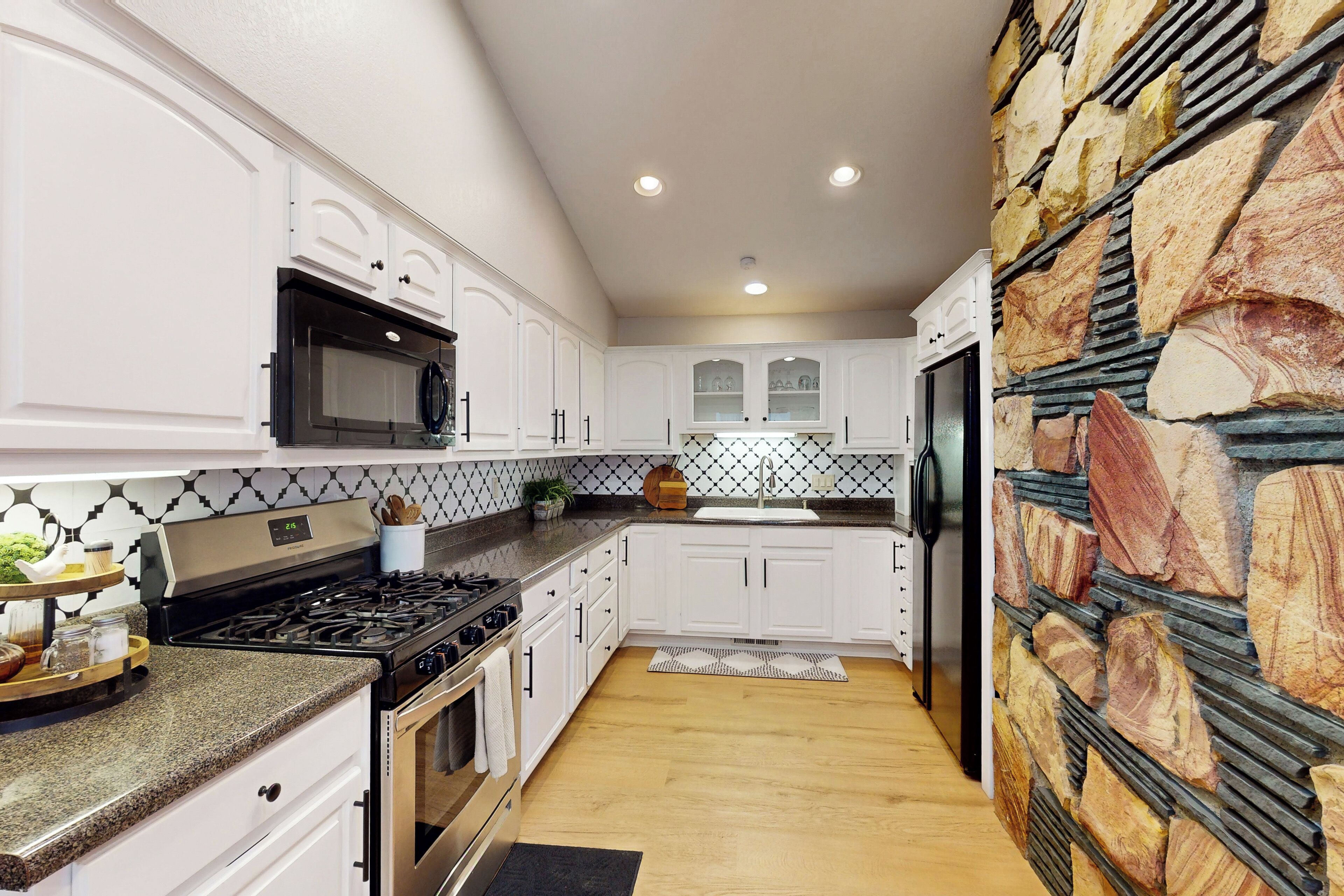 Uma cozinha moderna com armários brancos, bancada de granito, forno embutido e parede decorada com pedra.