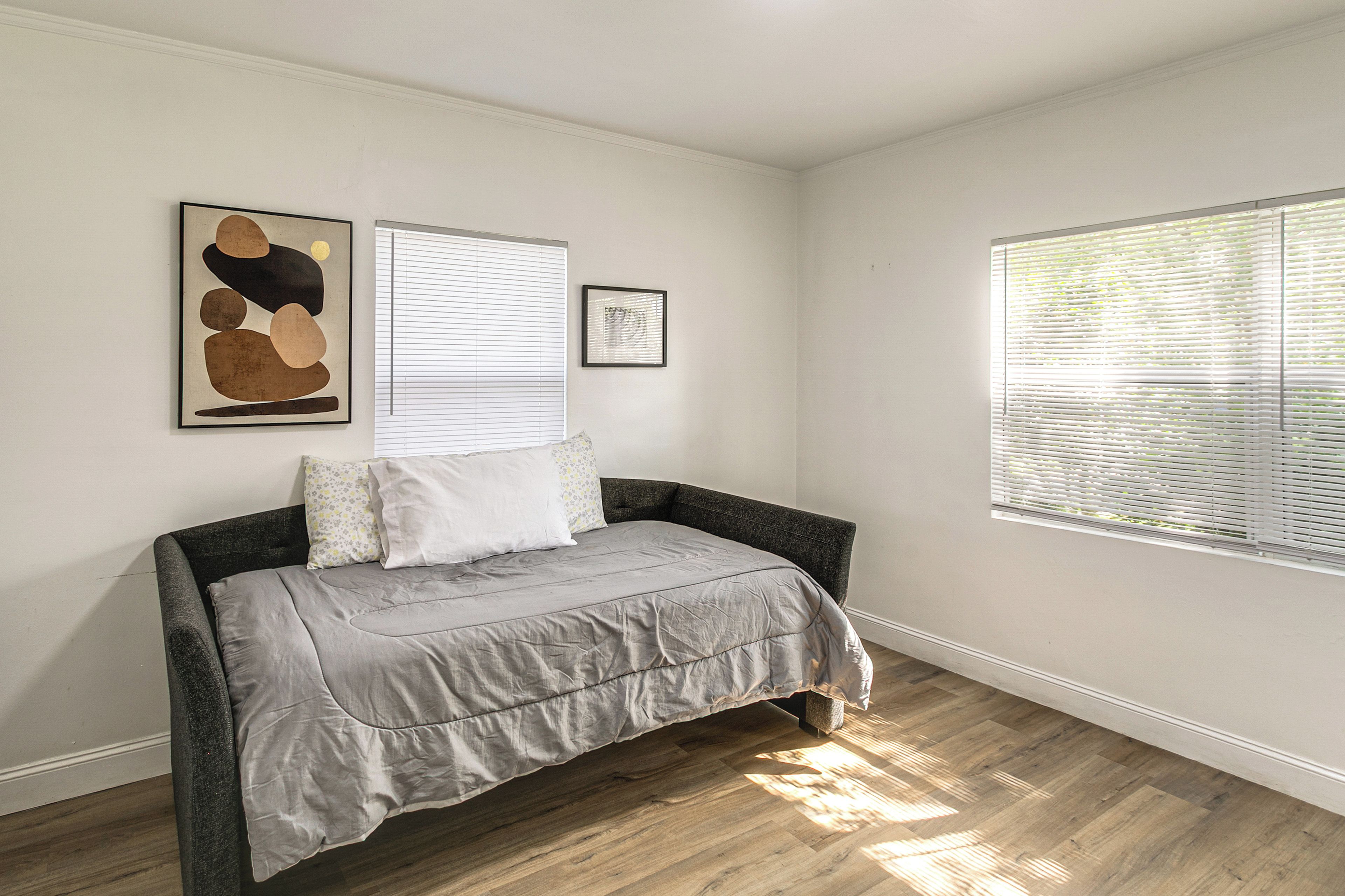 A bedroom with a bed, a window with blinds, and two framed artworks on the wall.