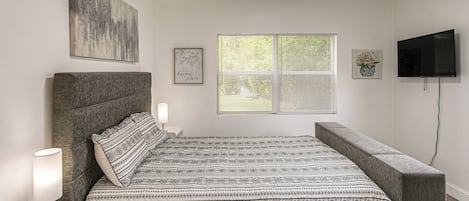 Un dormitorio con una cama, una mesita de noche, una lámpara, un televisor y una ventana con persianas.