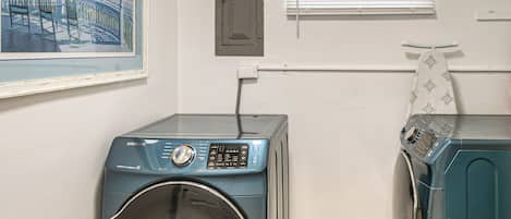 A laundry room with two front-loading washing machines, a door, a window with blinds, and a framed picture on the wall.