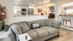 Ein modernes Wohnzimmer mit einem grauen Sofa, einer Fernbedienung auf einem Beistelltisch und einer Küche im Hintergrund.