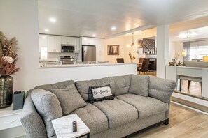 Una sala de estar moderna con un sofá gris, un control remoto sobre una mesita auxiliar y una cocina al fondo.