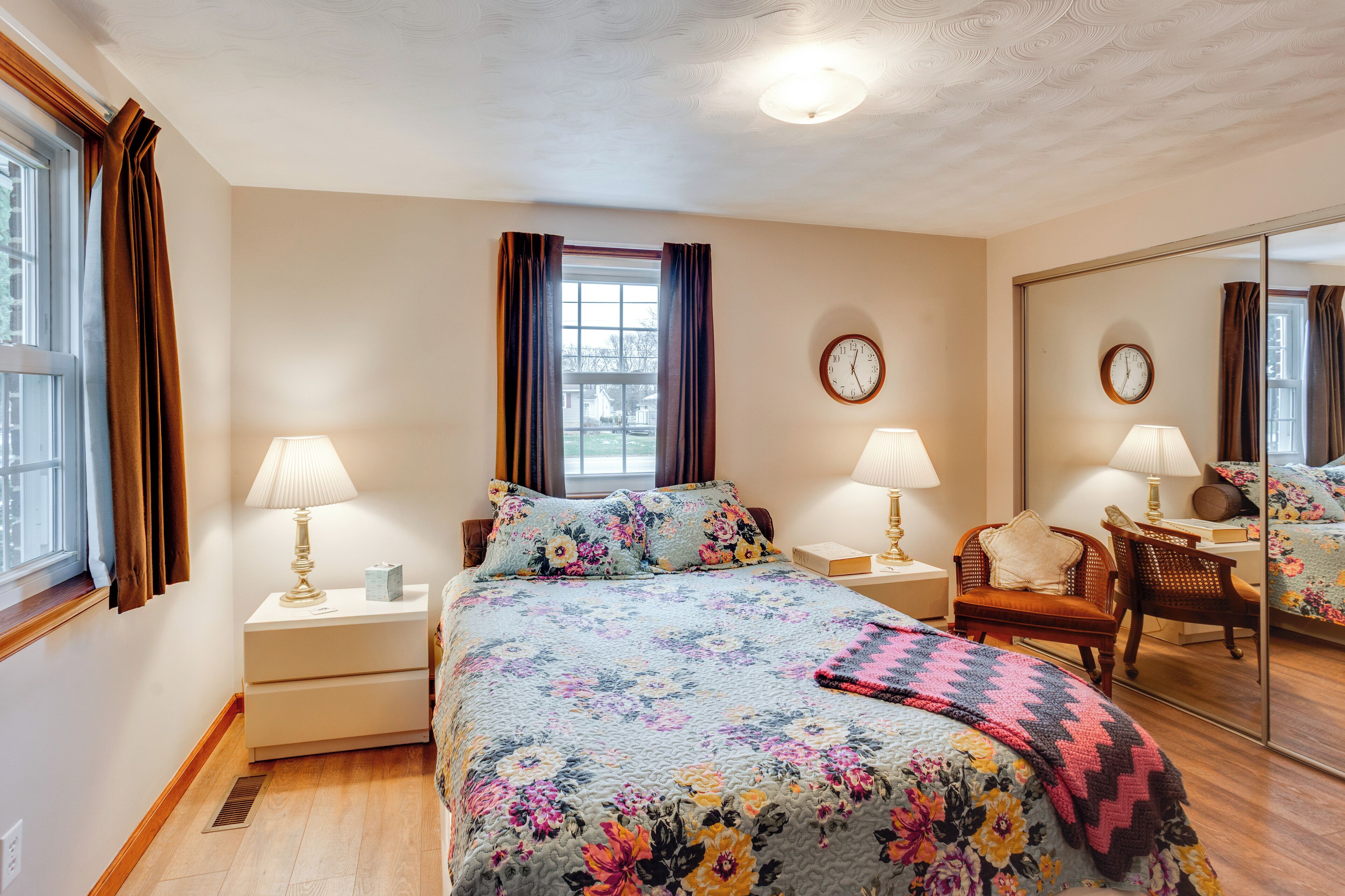 Quarto com colcha florida, duas luminárias de cabeceira, um espelho e uma janela com cortinas.