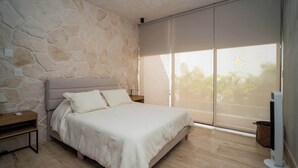 Una camera da letto con parete in pietra, un letto con biancheria bianca, un comodino, una lampada da comodino, un'ampia finestra con vista su palme e una ventola da terra.