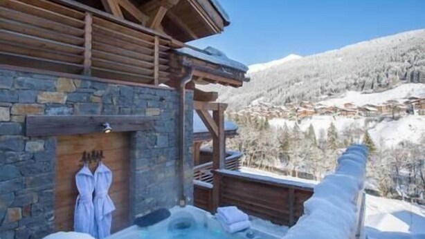 Una cabaña de madera con pared de piedra, un jacuzzi y vistas a un pueblo montañoso cubierto de nieve.