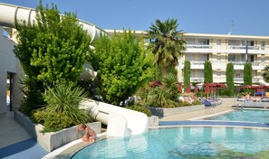 Area piscina con uno scivolo acquatico, immersa nel verde e con un edificio a più piani con balconi.