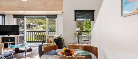 A modern living room with a glass dining table, leather chairs, a flat-screen TV, and a balcony with a view of the outdoors.