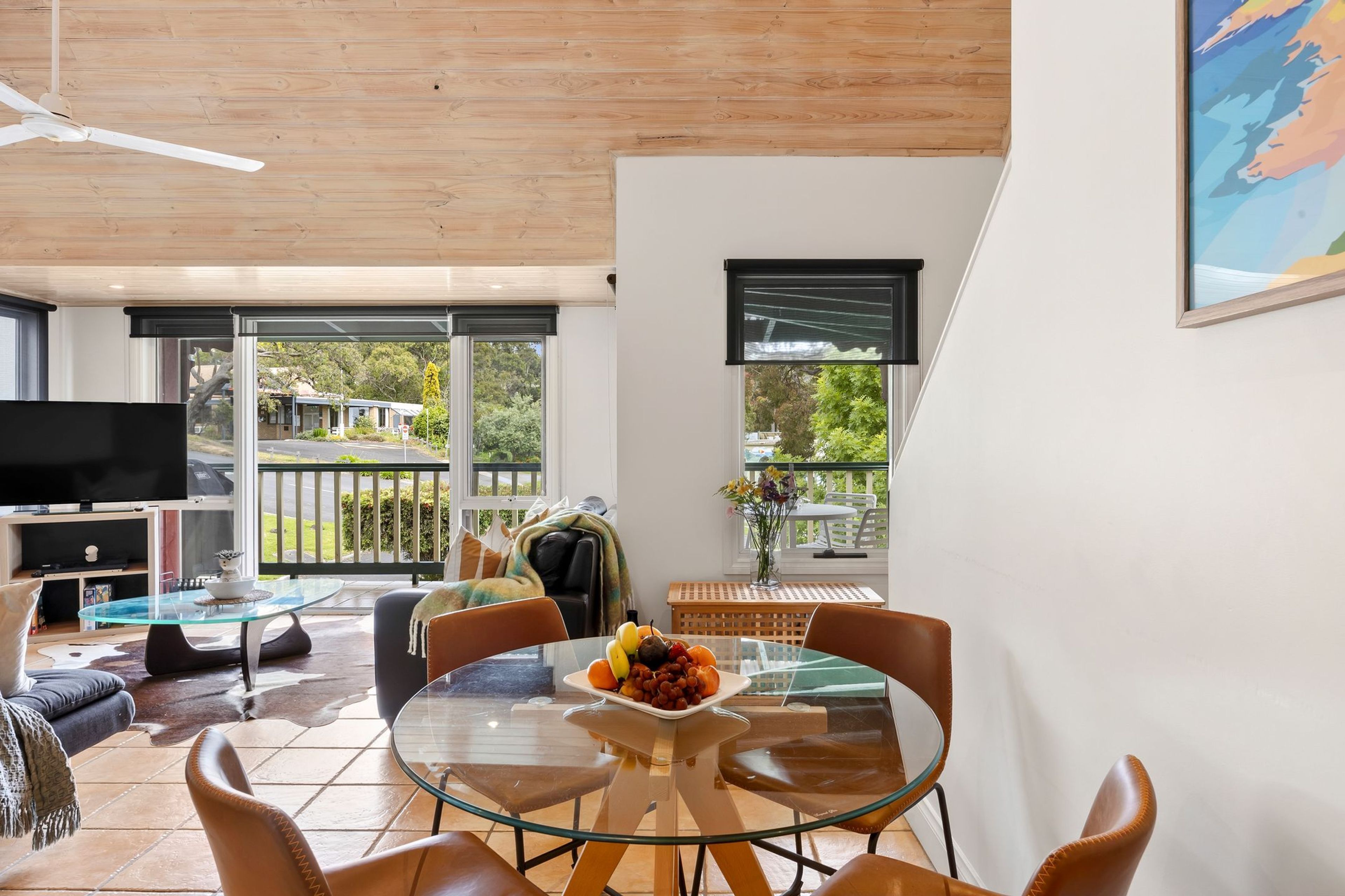 A modern living room with a glass dining table, leather chairs, a flat-screen TV, and a balcony with a view of the outdoors.