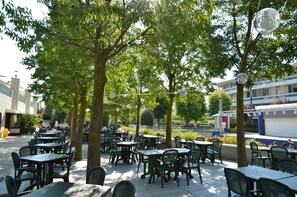 Een terras met tafels en stoelen onder bomen, omringd door gebouwen.