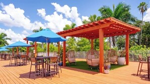 Un patio al aire libre con suelo de madera, sombrillas azules, sillas de mimbre y una pérgola roja.