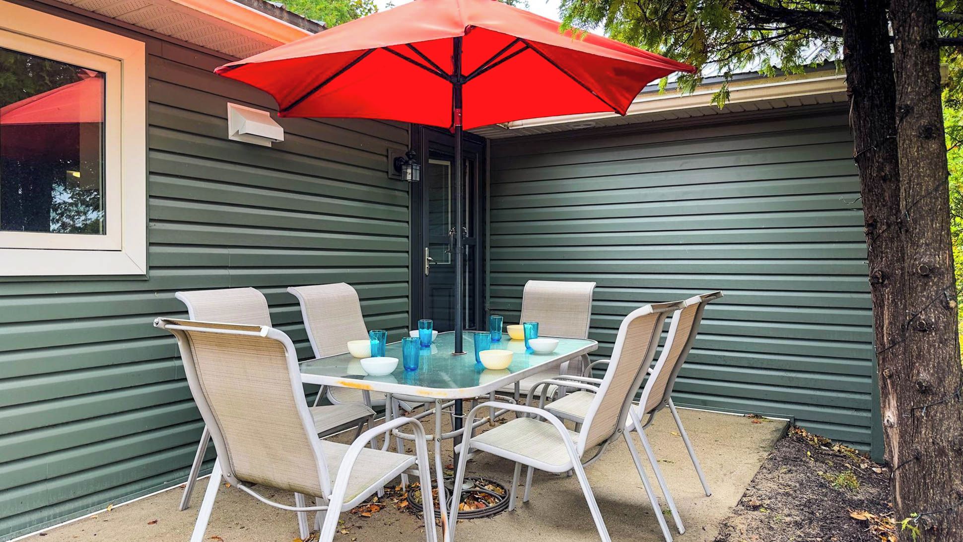 A patio with a table and chairs, a red umbrella, and a house with green siding.