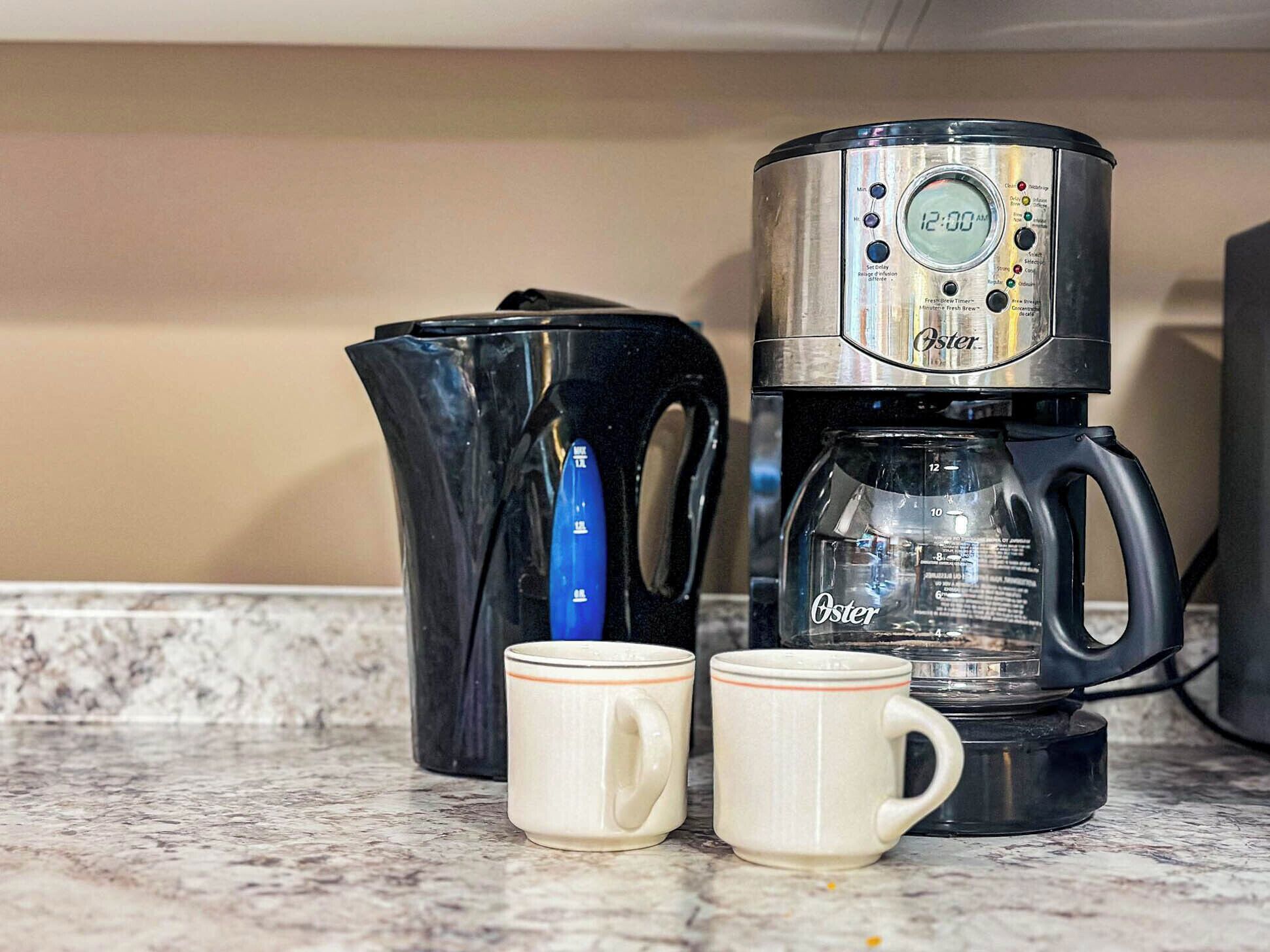 A coffee maker with a digital clock displaying 12:00 AM, two white mugs, and a black kettle on a kitchen counter.