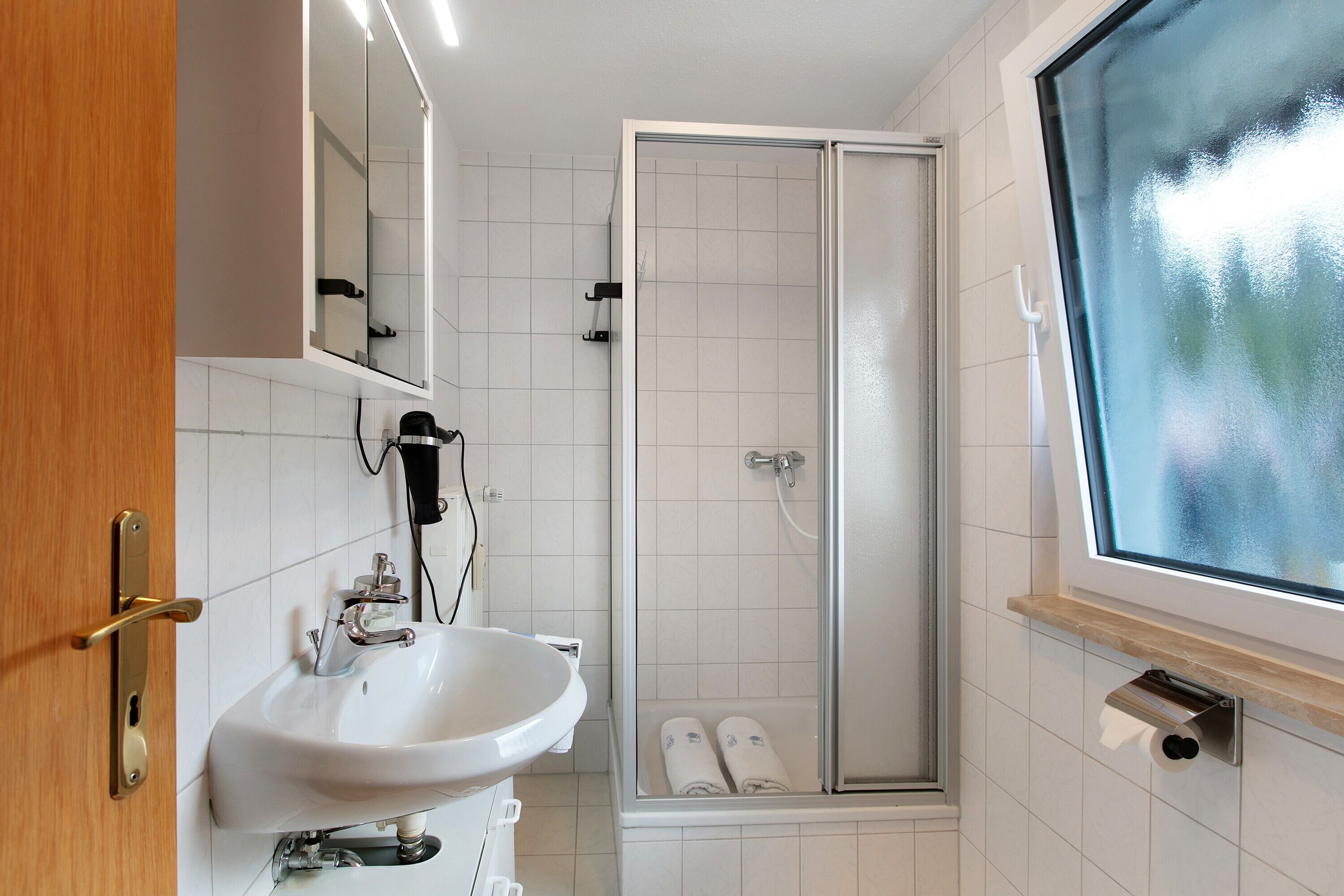 Ein Badezimmer mit einem weißen Waschbecken, einem Spiegel, einer Duschkabine mit Glastür und einem Fenster mit Ausblick.