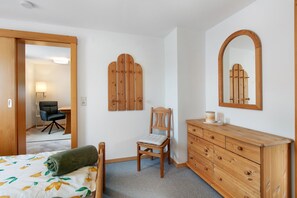 Un dormitorio con una cama, un aparador de madera, una silla, un espejo y una puerta que da a otra habitación.