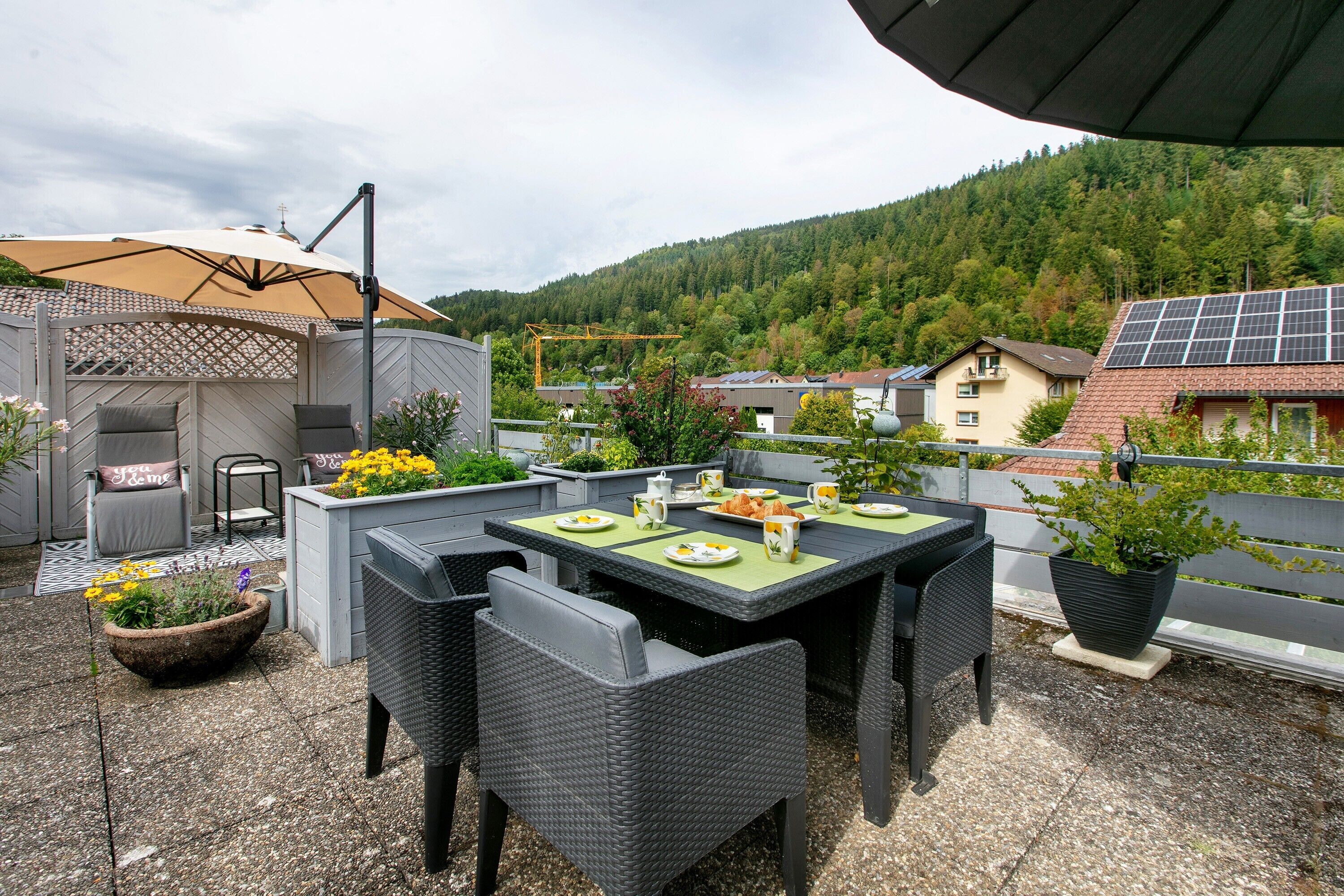 An outdoor patio with wicker furniture, a table set for a meal, and a view of a forested hillside.