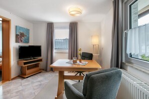 A hotel room with a flat-screen TV, a wooden TV stand, a dining table with a chair, a vase with flowers, a lamp, and a window with curtains. - Apartment in the Cathedral City, Haus Barbara 1st Floor with Mountain View, Terrace & Wi-Fi (St. Blasien)