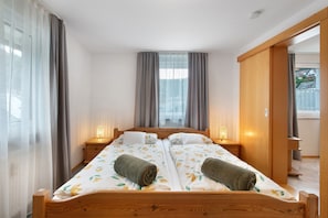 A double bed with a wooden headboard, two bedside tables with lamps, and a view of a residential area through the window. - Apartment in the Cathedral City, Haus Barbara 1st Floor with Mountain View, Terrace & Wi-Fi (St. Blasien)