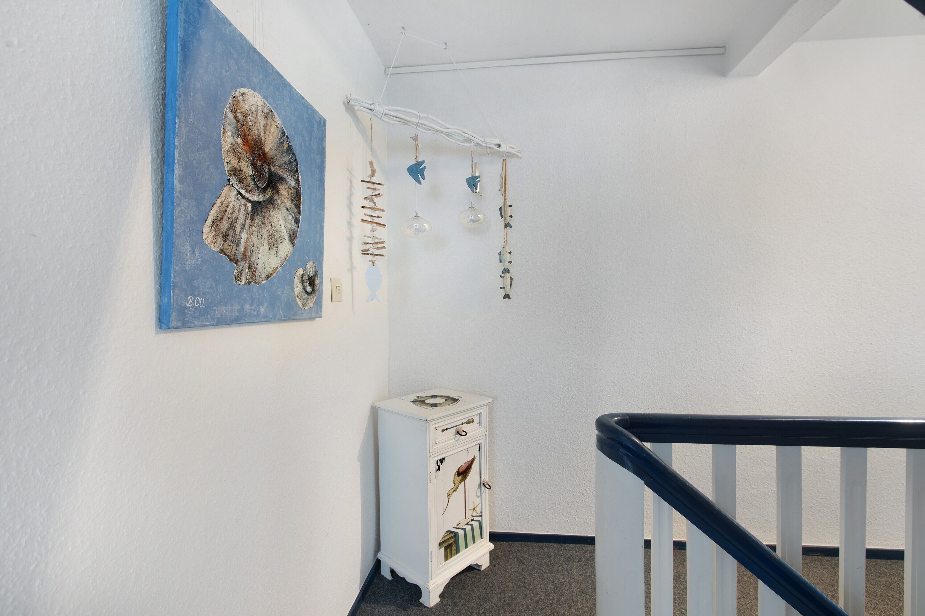 A hallway with a white wall, a blue painting, a small white cabinet, and a black railing.