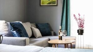 Moderni hotellihuone, jossa on harmaa sohva, pieni pyöreä pöytä, jonka päällä on pullo ja lasi, ruukkukasvi ja seinällä oleva kehystetty maisemamaalaus.