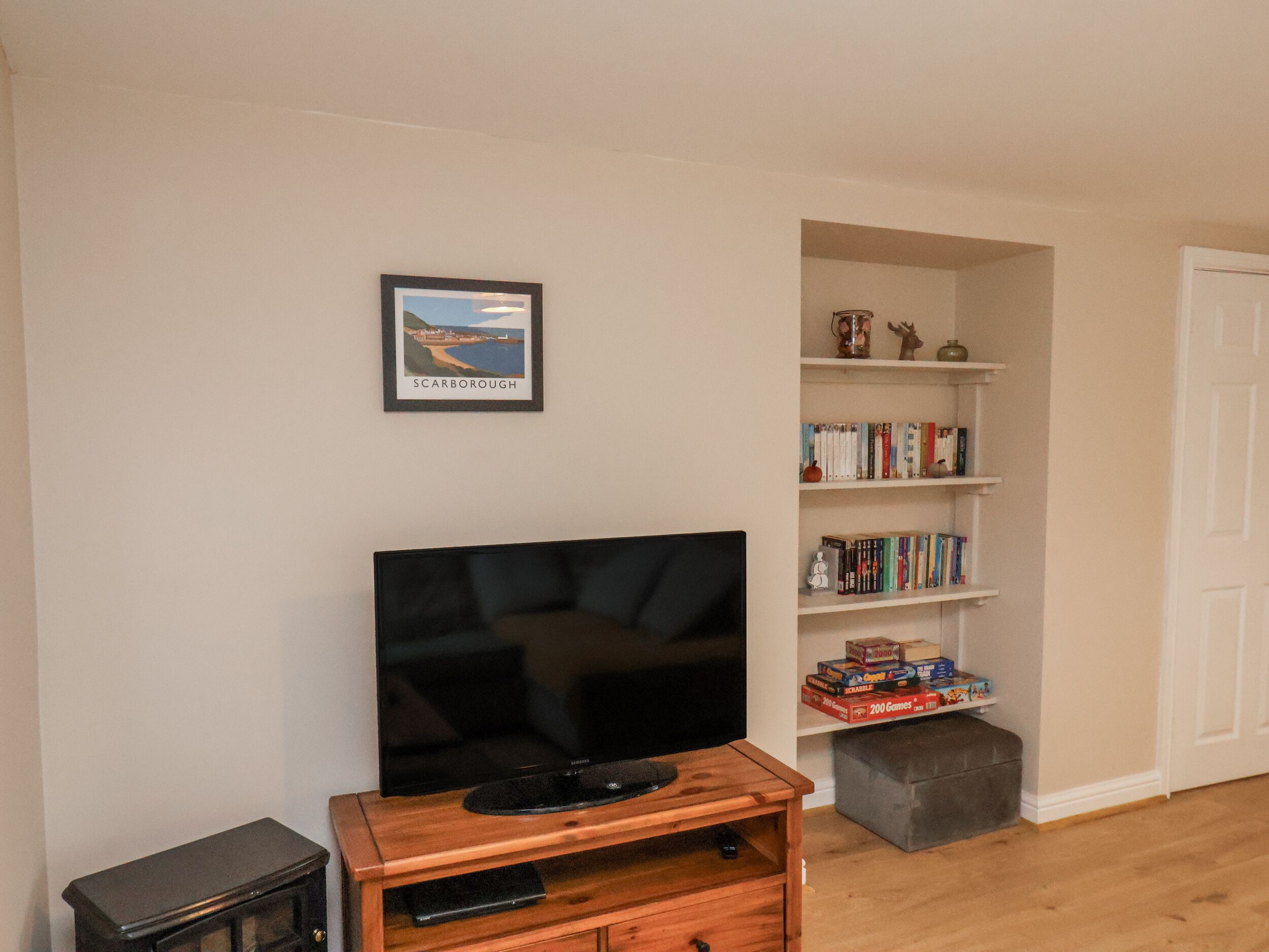 A living room with a flat-screen TV on a wooden stand, a bookshelf with books and board games, and a framed picture on the wall.