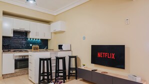 Eine moderne Küche mit weißen Schränken, einem eingebauten Herd und einer Mikrowelle. Es gibt einen Essbereich mit einem weißen Tisch und schwarzen Hocker. Ein großer Flachbildfernseher an der Wand zeigt das Wort "NETFLIX".