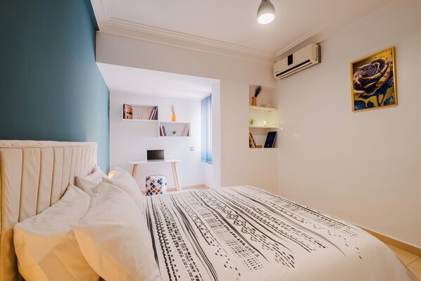 Una camera da letto con un letto, una scrivania, mensole piene di libri e oggetti decorativi, e un condizionatore d'aria.