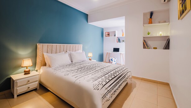 Ein Schlafzimmer mit Bett, Nachttischen, einer Lampe, einem Schreibtisch und Regalen mit Büchern und Dekoration.